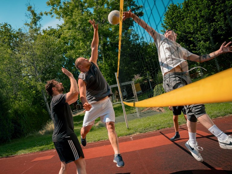 Drei Männer spielen Volleyball auf einem roten Außenplatz. Ein Spieler springt hoch, um den Ball über das Netz zu schlagen, während ein Gegenspieler versucht zu blocken. Im Hintergrund sind grüne Bäume und ein blauer Himmel zu sehen.