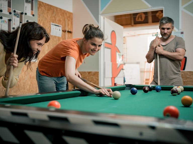 Zwei junge Frauen spielen Billard. Eine Frau im orangenen Shirt beugt sich zum Abstoß über den Tisch, während eine Freundin daneben steht und lächelnd zuschaut. Im Hintergrund beobachtet ein Mann das Spiel. Auf dem grünen Tuch liegen mehrere farbige Billardbälle.