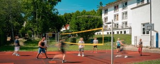 Eine Gruppe von Menschen spielt Volleyball auf einem roten Außenplatz. Ein gelbes Netz teilt das Spielfeld. Im Hintergrund sind grüne Bäume und ein weißes Gebäude unter blauem Himmel zu sehen.
