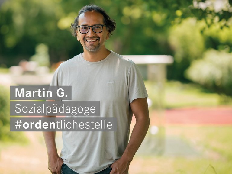 Ein lächelnder Mann mit Brille steht im Freien vor einem grünen Hintergrund. Ein Textfeld neben ihm identifiziert ihn als Martin G., Sozialpädagoge, und enthält den Hashtag #ordentlichestelle.