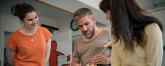 Drei Personen sitzen im Kreis. Ein Mann in der Mitte spielt Ukulele. Eine Frau links im orangen T-Shirt und eine Frau rechts im gelben Oberteil schauen ihm lächelnd zu.