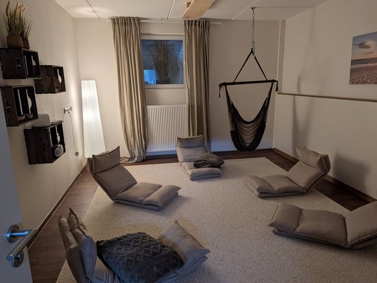 Ein entspannungsfreundlicher Raum mit mehreren grauen Bodensesseln, die kreisförmig auf einem Teppich angeordnet sind. Ein schwarzer Hängesessel hängt an der Decke. An der Wand sind Regale mit Pflanzen und Lautsprechern zu sehen.