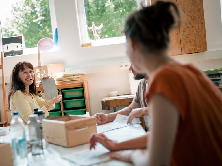 Eine glückliche Frau in einem gelben Oberteil sitzt an einem Tisch in einem hellen Atelier und hält eine Skizze hoch, um sie zwei anderen Personen zu zeigen. Im Hintergrund sind Staffeleien und Kunstmaterialien zu erkennen.