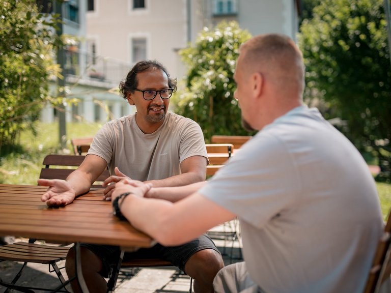 Zwei Männer unterhalten sich an einem Holztisch im Freien. Der Mann mit Brille und beigem T-Shirt gestikuliert beim Sprechen, während ihm sein Gesprächspartner zuhört. Im Hintergrund sind grüne Bäume und ein Haus zu sehen.