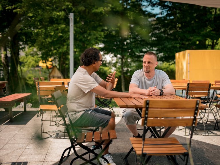 Zwei Männer sitzen auf einer Terrasse an einem Holztisch und unterhalten sich.