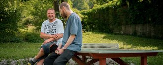 Zwei Männer sitzen lächelnd auf einem roten Tischtennistisch im Freien und unterhalten sich. Sie befinden sich in einer grünen Parkanlage mit Bäumen im Hintergrund.